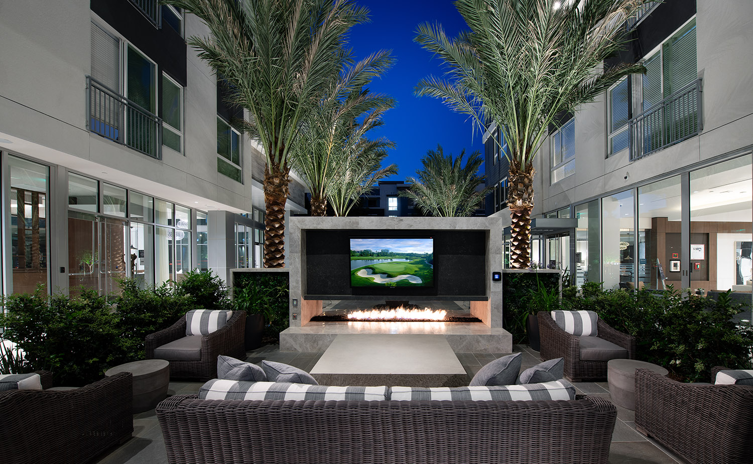 Outdoor fire put with flat screen TV and plush seating, surrounded by landscaping